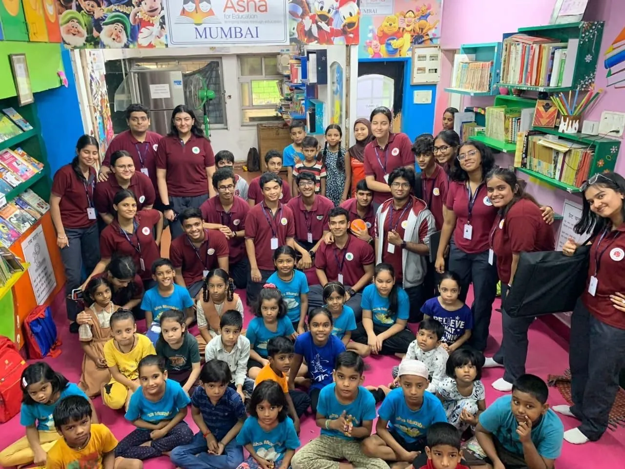 Group photo of volunteers in maroon shirts with children in blue T-shirts in a colourful classroom library