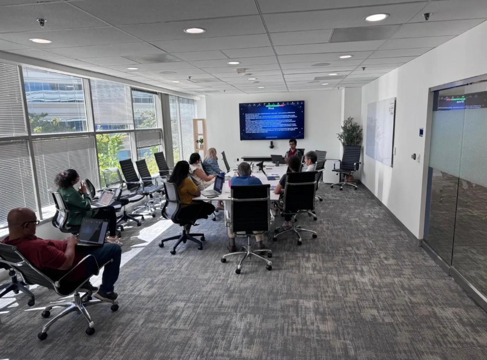 Aarav presenting market research to Celona’s team around a conference table, with the analysis on the wall screen behind him